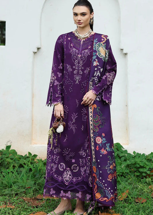 Woman wearing a long, dark purple embroidered dress with colorful patterns against a white wall.