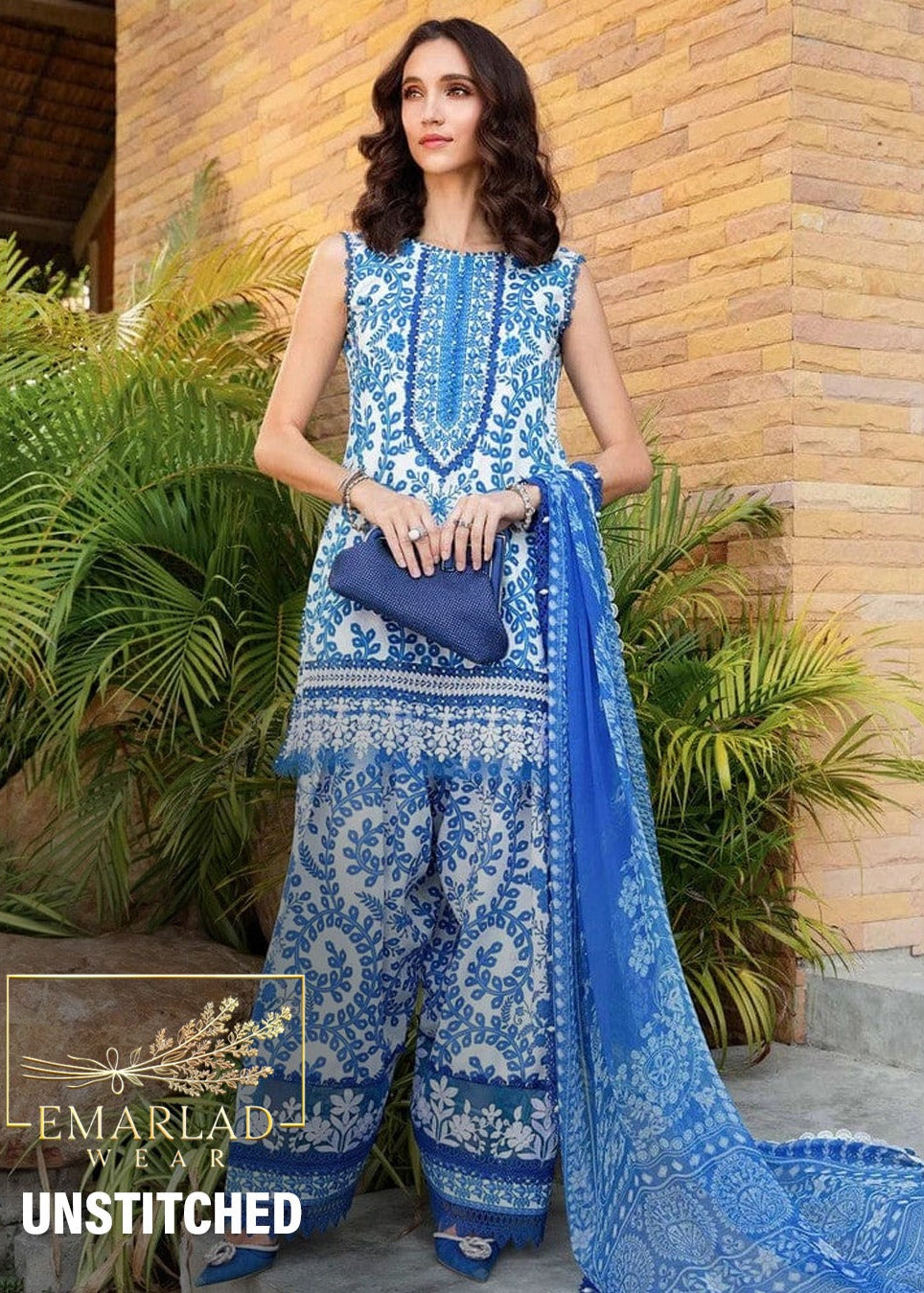 Woman wearing a blue and white patterned outfit with a blue dupatta, standing against a brick wall with plants.
